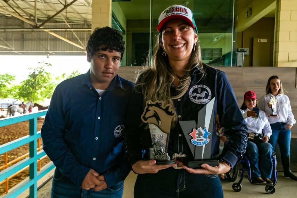 Descrição da imagem #PraCegoVer: O jovem Murilo com a equoterapeuta Eliane Baatsch. Murilo é um rapaz moreno, de cabelos pretos. Ele veste camisa azul marinho. Eliane é uma mulher morena, de cabelos lisos e compridos. Ela está usando camisa azul e um boné. Eliane segura o troféu de Murilo. Fim da descrição.