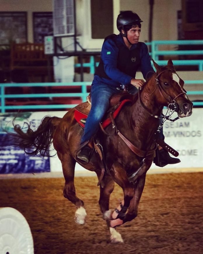 Descrição da imagem #PraCegoVer: O jovem Murilo durante torneio equestre. Murilo é um rapaz moreno e está montado em um cavalo marrom. Murilo veste calça jeans e camisa azul marinho. Ele está usando capacete. Fim da descrição.