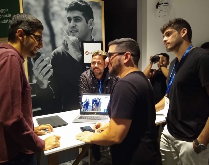 Descrição da imagem #PraCegoVer: Sundar Pichai, Thadeu Luz, Ronaldo Tenório e Carlos Wanderlan estão em volta de uma mesa com um notebook aberto mostrando o trabalho da tradução em Língua de Sinais. No fundo existe um quadro em preto e branco com a foto de Ronaldo Tenório e ao lado desse quadro um fotógrafo registrando o momento. Fim da descrição.