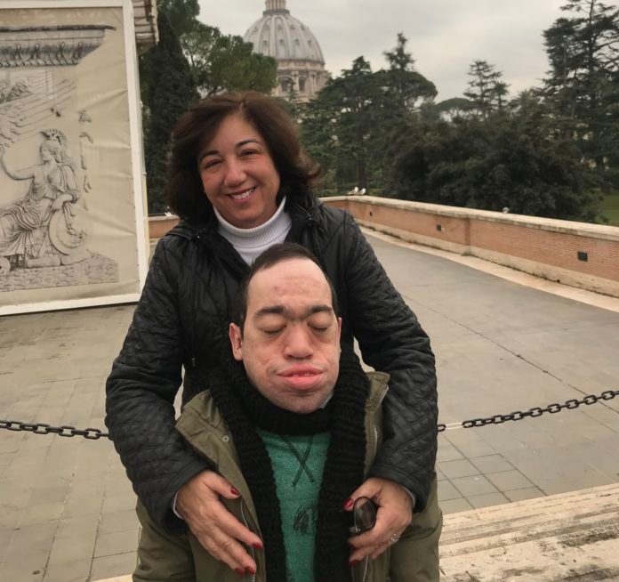 descrição da imagem #PraCegoVer: Regina Próspero, vice-presidente do Instituto Vidas Raras, está com seu filho, Dudu Próspero. Regina está atrás de Dudu, e o abraça. Regina é uma mulher morena, de pele clara. Ela tem cabelos pretos, lisos, na altura dos ombros. Dudu é um rapaz moreno, de pele clara e cabelos castanhos. Dudu tem mucopolissacaridose. Fim da descrição.