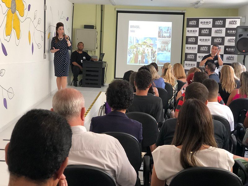 Descrição da imagem #PraCegoVer: Sala do curso de moda inclusiva. Várias pessoas estão sentadas e assistem a aula. Uma mulher está à frente do grupo e ao seu lado está um tradutor de Libras. Fim da descrição.