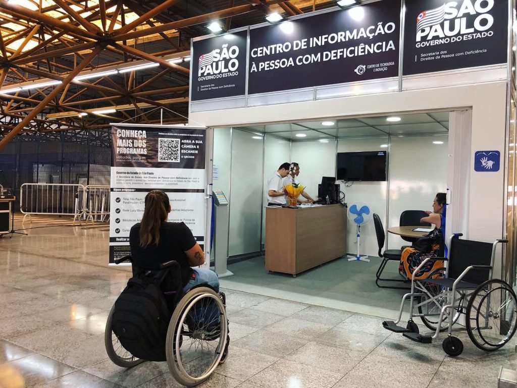 Descrição da imagem #pracegover: Foto do espaço do centro de informação para pessoas com deficiência na estação Tatuapé. Na parte de fora está uma mulher em sua cadeira de rodas. Dentro estão pessoas em pé atrás de um balcão e também tem uma pessoa sentada. Fim da descrição.