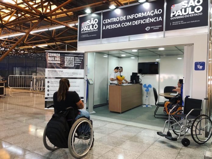 Descrição da imagem #pracegover: Foto do espaço do centro de informação para pessoas com deficiência na estação Tatuapé. Na parte de fora está uma mulher em sua cadeira de rodas. Dentro estão pessoas em pé atrás de um balcão e também tem uma pessoa sentada. Fim da descrição.