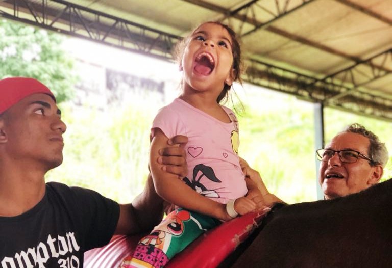 Descrição da imagem #PraCegoVer: Uma menina no cavalo durante equoterapia. Ela está sentada no dorso do cavalo e ao seu lado estão dois profissionais de equoterapia. Ela tem paralisia cerebral e está sorrindo. Fim da descrição.