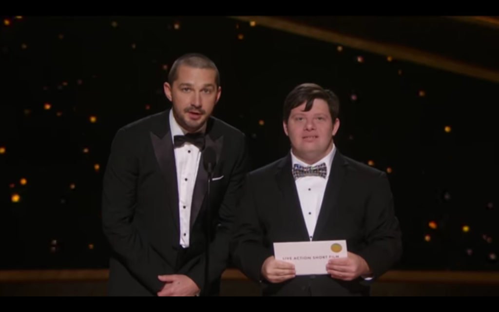 Descrição da imagem #PraCegoVer: Os atores Shia LaBeouf e Zack Gottsagen no palco do Oscar 2020. Shia e Zack são homens morenos, de pele clara e cabelos castanhos. Zack tem síndrome de Down. Eles usam traje de gala e gravata borboleta. Zack está segurando o envelope com o nome do vencedor na categoria. Fim da descrição.