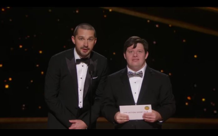 Descrição da imagem #PraCegoVer: Os atores Shia LaBeouf e Zack Gottsagen no palco do Oscar 2020. Shia e Zack são homens morenos, de pele clara e cabelos castanhos. Zack tem síndrome de Down. Eles usam traje de gala e gravata borboleta. Zack está segurando o envelope com o nome do vencedor na categoria. Fim da descrição.