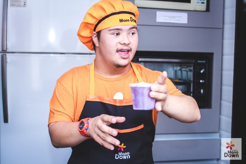 Descrição da imagem #PraCegoVer: Um jovem com síndrome de Down está usando um uniforme laranja e segura um suco com a mão esquerda. Fim da descrição.