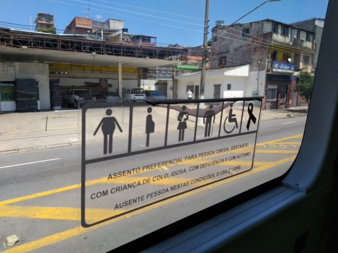 Descrição da imagem #PraCegoVer: A janela de um ônibus com o novo adesivo dos assentos preferenciais, com o símbolo do autismo. Fim da descrição.