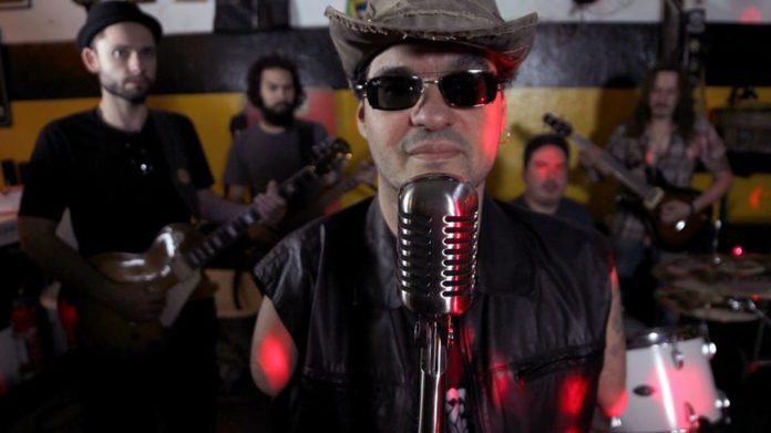 Descrição da imagem #PraCegoVer: Dudé está à frente de sua banda. Ele é um homem moreno, de pele clara. Ele está usando um chapeu e óculos escuros. Fim da descrição.