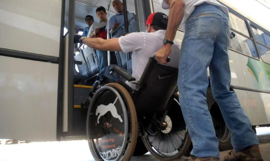 Descrição da imagem #PraCegoVer: Um homem cadeirante está entrando em um ônibus. Um homem está ajudando, conduzindo a cadeira de rodas. Fim da descrição.