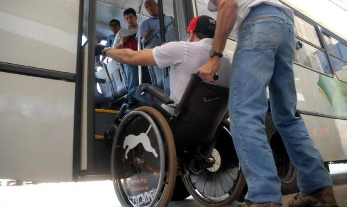 Descrição da imagem #PraCegoVer: Um homem cadeirante está entrando em um ônibus. Um homem está ajudando, conduzindo a cadeira de rodas. Fim da descrição.