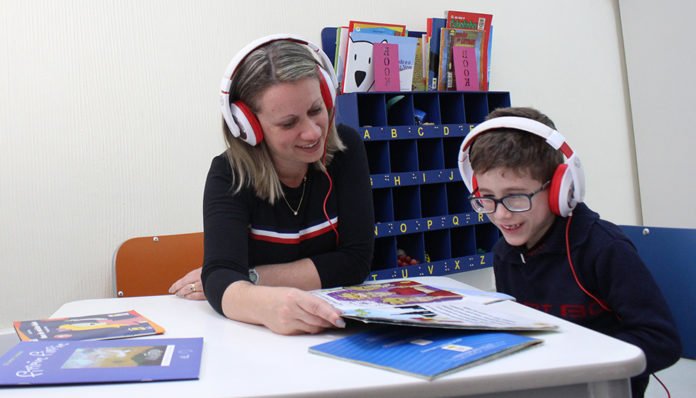 #descriçãodaimagem: Foto de um menino e uma mulher. Eles estão sentados em uma mesa. À sua frente tem um livro aberto e ambos usam fones de ouvido. Eles estão fazendo a leitura de um audiolivro. Fim da descrição.