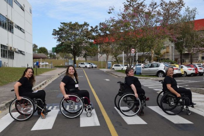 Descrição da imagem #PraCegoVer: As quatro consultoras da Talento Incluir estão atravessando a rua, na faixa de pedestres. Elas são cadeirantes e estão vestindo roupas pretas. Fim da descrição.