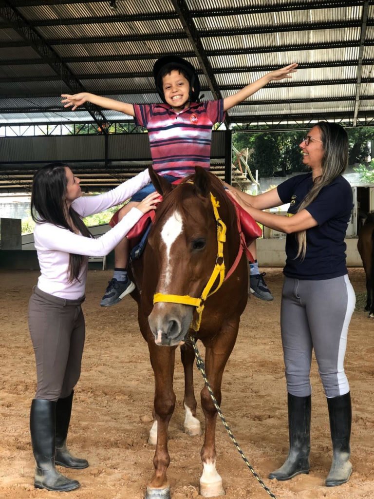 Descrição da imagem #PraCegoVer: Um menino está montado no cavalo. Ele está com os braços abertos. Ao lado do cavalo tem duas profissionais de equoterapia. Fim da descrição.