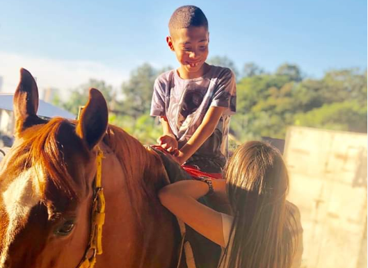 Descrição da imagem #PraCegoVer: Um menino está montado no cavalo. Uma equoterapeuta está ao lado do cavalo. Ela conversa e auxilia o menino. Fim da descrição.