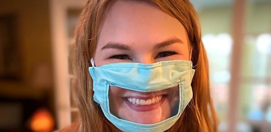 Descrição da imagem #PraCegoVer: A estudante Ashley usando uma máscara adaptada, que é azul clara e tem uma abertura transparente na parte da boca. Ashley é uma moça loira, de cabelos compridos e está sorrindo. Fim da descrição.