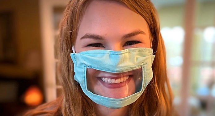 Descrição da imagem #PraCegoVer: A estudante Ashley usando uma máscara adaptada, que é azul clara e tem uma abertura transparente na parte da boca. Ashley é uma moça loira, de cabelos compridos e está sorrindo. Fim da descrição.
