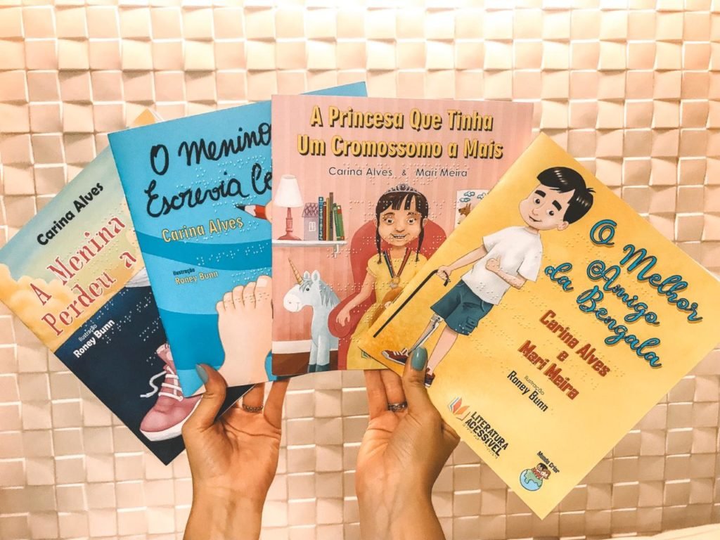 descrição da imagem #PraCegoVer: Uma mão está segurando quatro livros infantis. Fim da descrição.