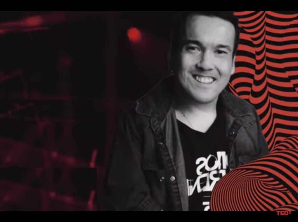 Descrição da imagem #PraCegoVer: O humorista Gigante Leo em arte de divulgação do TEDx. Leonardo é um homem branco, de cabelos pretos e ele está sorrindo. O fundo da imagem é preto e na parte lateral direita da imagem tem algumas listras onduladas vermelhas. Fim da descrição.