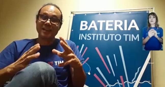 Descrição da imagem #pracegover: O professor de bateria do Instituto TIM. Ele está sentado e atrás dele tem um banner escrito: Bateria Instituto TIM. No canto superior direito tem a janela de tradução em Libras, e nela está uma intérprete. Fim da descrição.