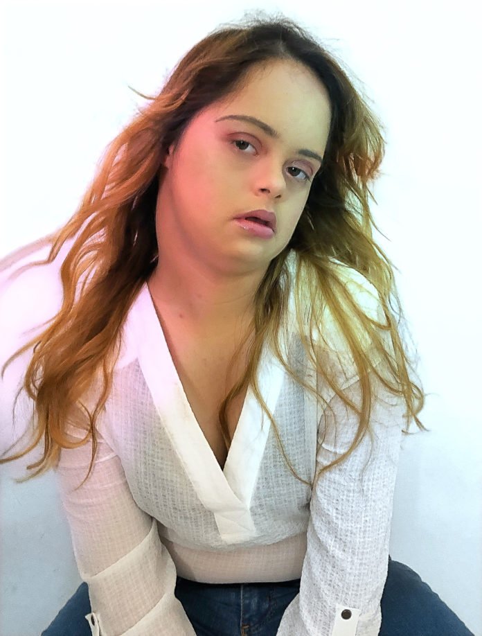 Descrição da imagem #pracegover: A influenciadora Cacai Bauer. Ela é morena, tem pele clara, e cabelos longos e loiros. Ela está usando uma camisa branca e faz pose para a foto. Cacai tem síndrome de Down. Fim da descrição.