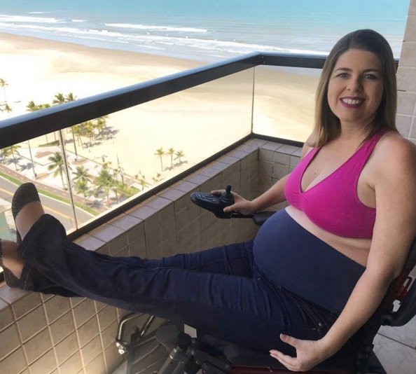 Descrição da imagem #pracegover: Ketly Vieira está na varanda de um apartamento à beira mar. Ketly está grávida. Ela usa um top pink e uma calça preta. Fim da descrição.