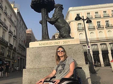 Descrição da imagem #pracegover: Ketly Vieira está em um marco na cidade de Madri, na Espanha. Ketly tem pele clara e cabelos lisos e loiros, um pouco abaixo dos ombros. Ela usa uma blusa cinza. Ketly está em sua cadeira de rodas e sorri. Fim da descrição.