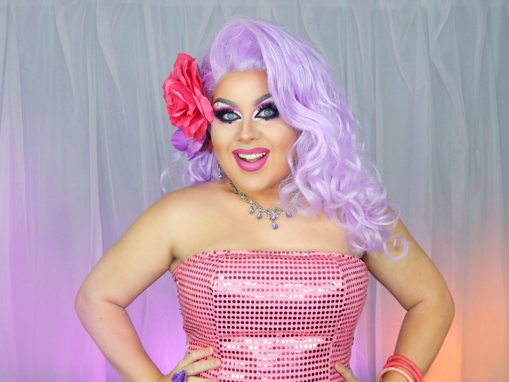 Descrição da imagem #pracegover: Foto da influenciadora digital Kitana Dreams. Ela é uma Drag Queen, branca, tem cabelos compridos lilás. Kitana está usando um tomara que caia pink de paetês e tem uma flor pink nos cabelos. Fim da descrição.