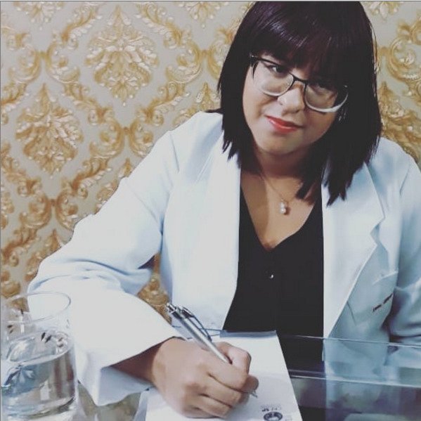 Descrição da imagem #pracegover: A doutora Grayce França, primeira farmacêutica com deficiência visual de São Paulo. Ela é uma mulher morena, de pele clara. Tem cabelos pretos, lisos, na altura dos ombros e usa uma franja. Grace está sentada à frente de uma mesa, onde está um papel onde ela escreve. Ela usa uma blusa preta e um jaleco branco. Grayce usa óculos de grau e está olhando em direção à câmera fotográfica. Ao fundo, a parede tem papel de parede estilizado cor de ouro. Fim da descrição.
