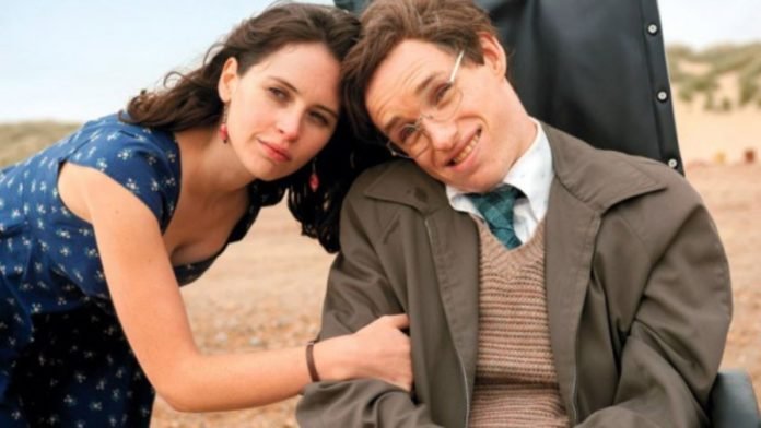 Descrição da imagem #pracegover: Cena do filme A teoria de tudo. Na imagem vemos os atores que interpretam Stephen Hawking e sua esposa Jane Wilde. Ele está em sua cadeira de rodas e ela está ao seu lado, segurando em seu braço. Fim da descrição.