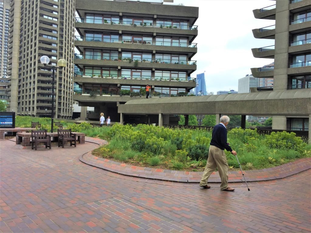 Descrição da imagem #pratodosverem: Um idoso caminha na calçada. Ele usa uma bengala e ao fundo vemos um local organizado e arborizado. Fim da descrição. do governo francês. Fim da descrição.