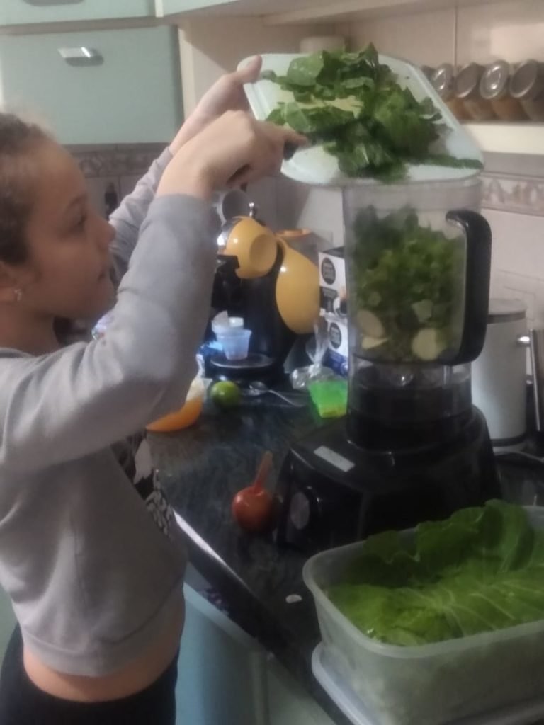 Descrição da imagem #pratodosverem: Uma menina está preparando um suco verde. Ela coloca os ingredientes no liquidificador. Fim da descrição.