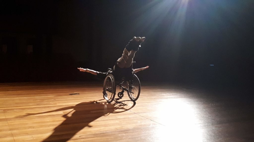 Descrição da imagem #pracegover: Uma apresentação da cia de dança. O dançarino está no palco e faz uma coreografia na cadeira de rodas. Fim da descrição.