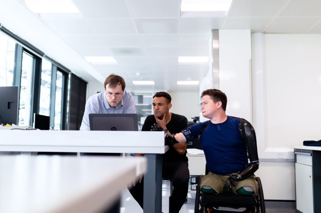 Descrição da imagem #pracegover: Três homens estão em um escritório. Um deles tem deficiência física. Ele tem amputação bilateral de braços e pernas. Eles estão vendo coisas em um computador. Fim da descrição.