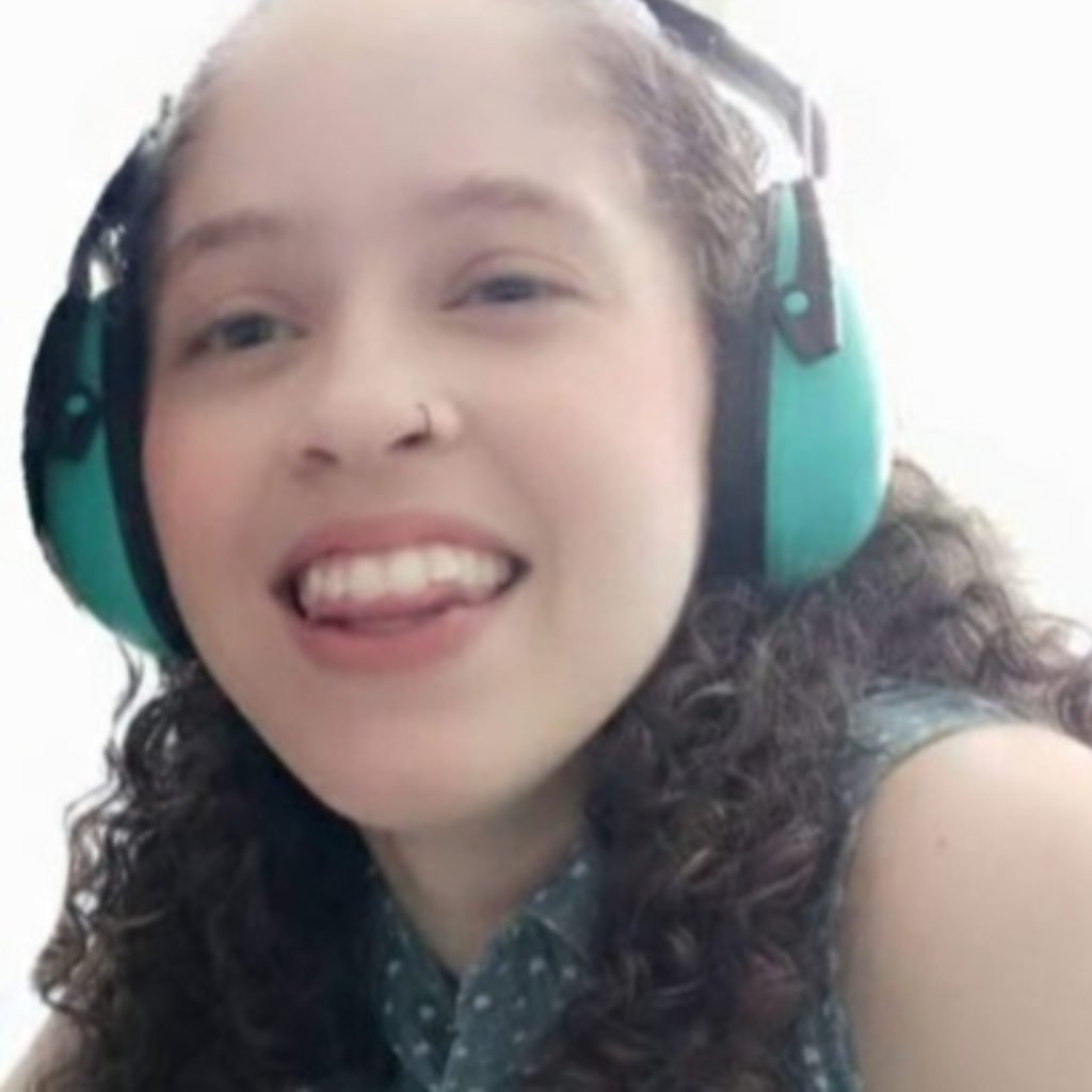 Descrição da imagem #pracegover: Uma foto de Monique. Ela é uma moça morena de cabelos cacheados, castanhos. Ela está sorrindo e tem um fone de ouvidos na cabeça. Fim da descrição.