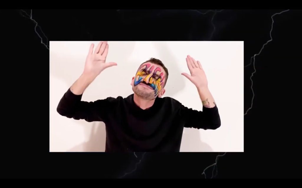 Descrição da imagem #pratodosverem: Cena do clipe em Libras da música Rajadão. Thyago Santos está com as mãos para cima. Ele faz o sinal de 'rajadão'. Ele usa camisa preta e está com o rosto pintado. Fim da descrição.