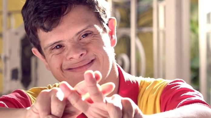 Descrição da imagem #pracegover: Close de um rapaz, com síndrome de Down. Ele está fazendo o sinal de hashtag com os dedos da mão, e sorri. Ele é moreno, tem cabelos castanhos, lisos e curtos. Fim da descrição.