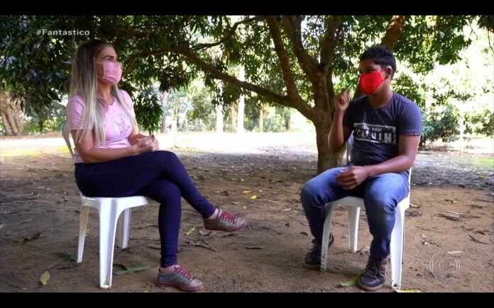 Descrição da imagem #pracegover: A professora Joyce e o aluno surdo Edilson, durante a aula. Eles estão sentados embaixo de uma árvore. Fim da descrição.