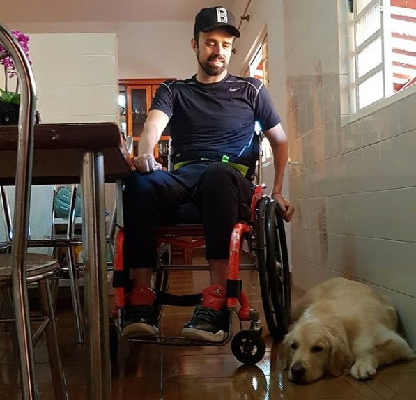 Descrição da imagem #pratodosverem: Bruno em sua casa. Ao seu lado está Kira, seu cão de assistência, que está deitada no chão. Fim da descrição.