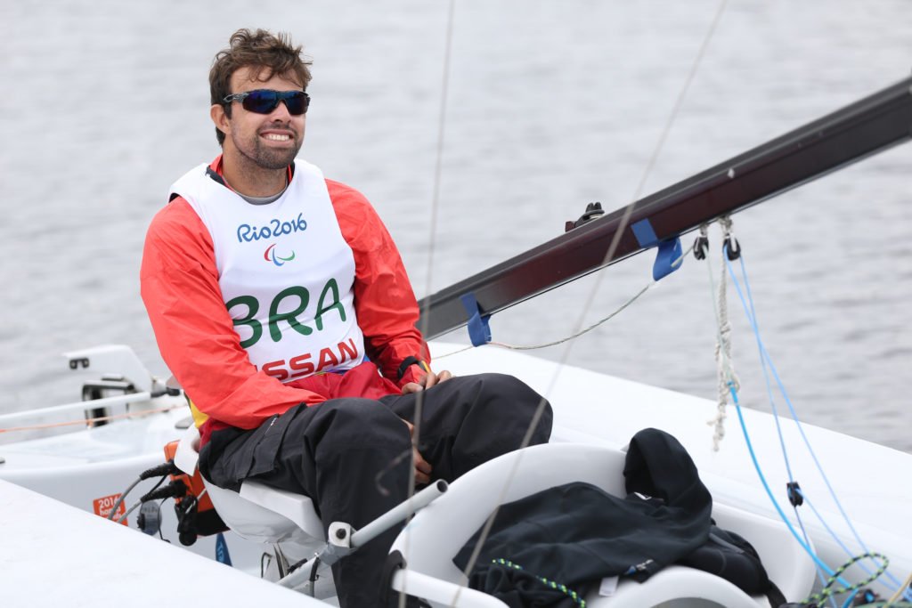 Descrição da imagem #pratodosverem: Foto do Bruno durante as Paralimpíadas do Rio de Janeiro. Ele está sentado no barco, durante treino na Marina da Glória. Bruno é um homem moreno, de pele clara e cabelos castanhos, curtos. Ele usa uma blusa vermelha, uma calça preta e um colete branco, onde está escrito: Rio2016 - BRA. Fim da descrição.