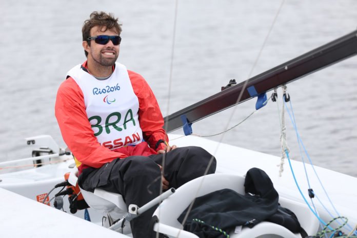 Descrição da imagem #pratodosverem: Foto do Bruno durante as Paralimpíadas do Rio de Janeiro. Ele está sentado no barco, durante treino na Marina da Glória. Bruno é um homem moreno, de pele clara e cabelos castanhos, curtos. Ele usa uma blusa vermelha, uma calça preta e um colete branco, onde está escrito: Rio2016 - BRA. Fim da descrição.