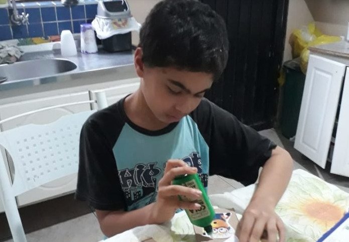 Descrição da imagem #pratodosverem: Um menino está fazedo um trabalho de colagem. Ele é moreno e tem cabelos pretos. Fim da descrição.