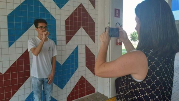 Descrição da imagem #pratodosverem: Um rapaz faz sinais em Libras. Ele está sendo filmado por uma mulher. Fim da descrição.