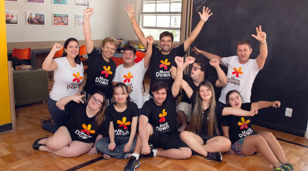 Descrição da imagem #pracegover: Leonardo Gontijo e Dudu do Cavaco com atendidos pelo Instituto Mano Down. Todos estão usando a camiseta do instituto. Eles estão sentados no chão da Casa Modelo. Fim da descrição.