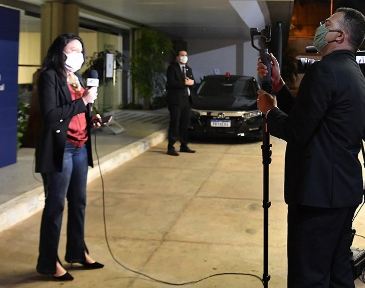 Descrição da imagem #pratodosverem: Uma repórter durante uma transmissão ao vivo. Ela está usando uma máscara branca. Fim da descrição.