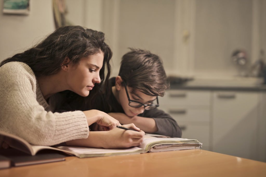 Descrição da imagem #pratodosverem: Uma mulher está ao auxiliando um menino durante os estudos. Fim da descrição.