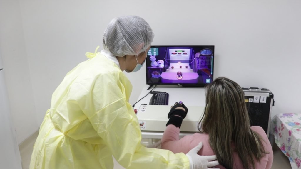 Descrição da imagem #pratodosverem: Uma mulher está realizando uma terapia em um equipamento. Ela está sendo auxiliada por uma profissional da saúde. Fim da descrição.
