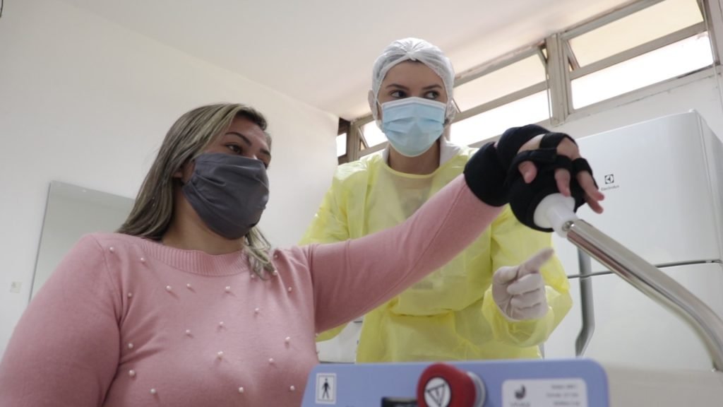 Descrição da imagem #pratodosverem: Uma mulher está realizando uma terapia em um equipamento. Ela está sendo auxiliada por uma profissional da saúde. Fim da descrição.