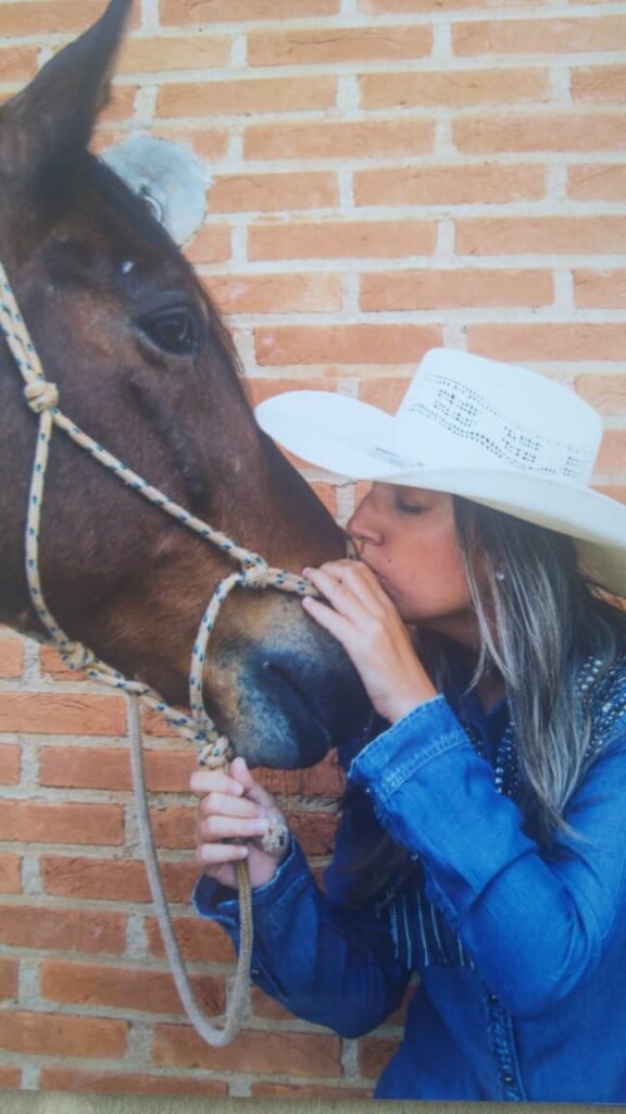 Descrição da imagem #pracegover: Veri está beijando um cavalo marrom. Ela é uma moça morena e está usando camisa jeans e chapéu branco. Fim da descrição.