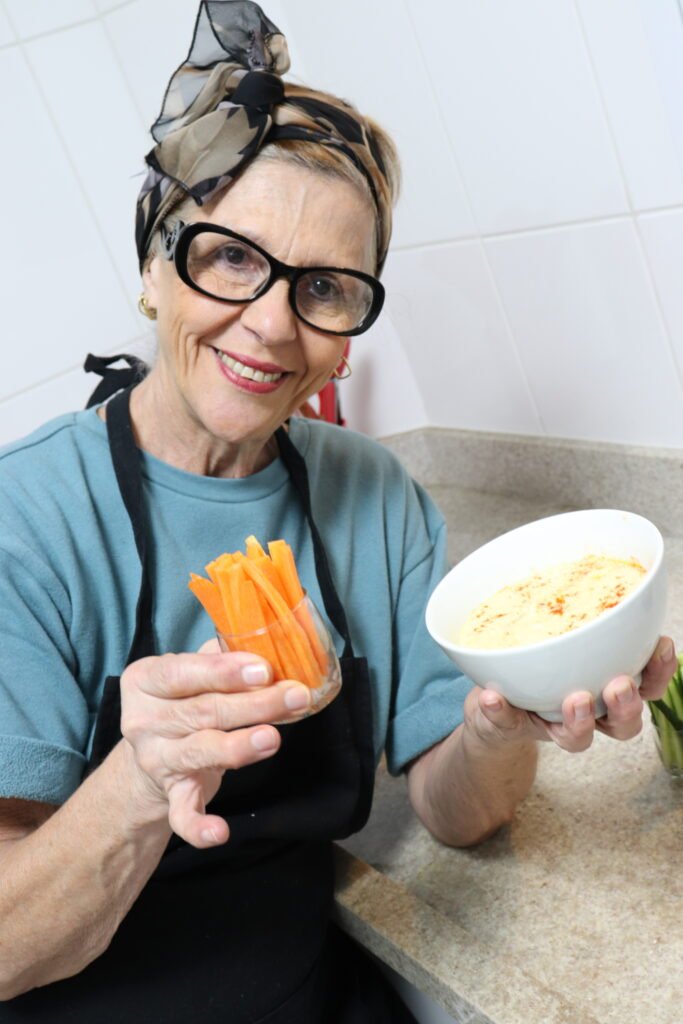 Descrição da imagem #pracegover: Foto da Vera, idealizadora do canal Cozinhando em Libras. Vera está na cozinha. Ela segura uma travessa com patê e um copo com palitinhos de cenoura. Vera é uma mulher morena. Ela usa óculos e está usando uma blusa azul, um avental preto e uma bandana nos cabelos. Fim da descrição.